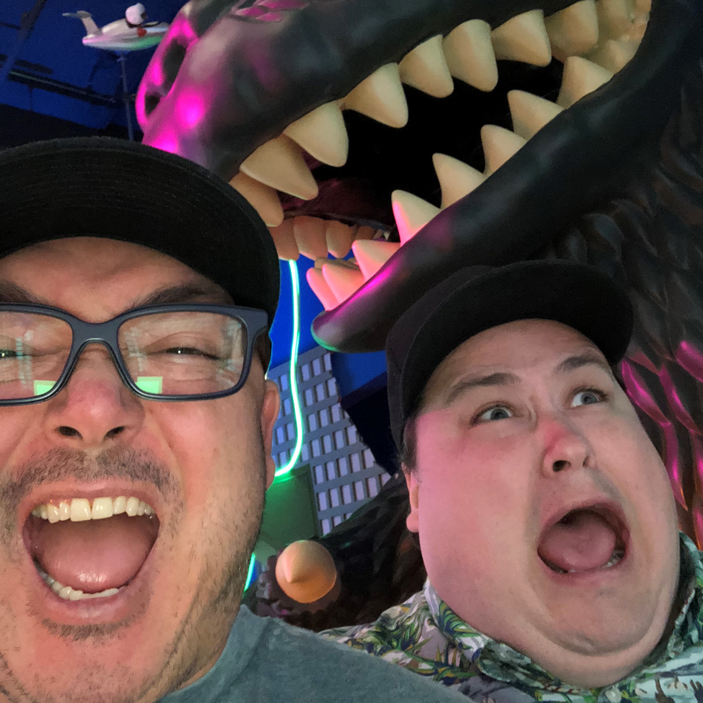two terrified mens faces who apear to be fleaing from a Godzilla statue towering above them
